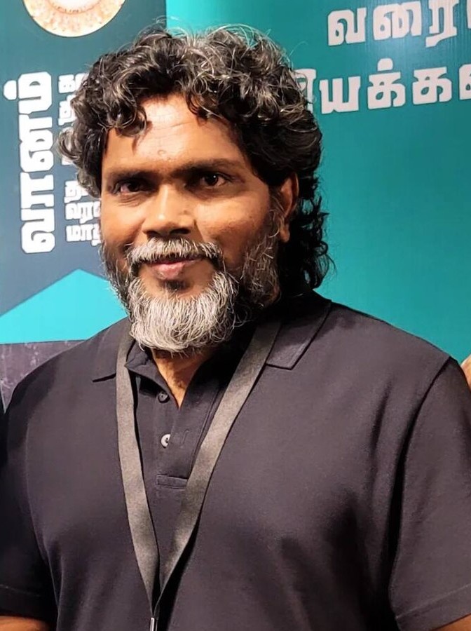 Pa. Ranjith in a black shirt and festival lanyard, in front of a Vaanam festival banner